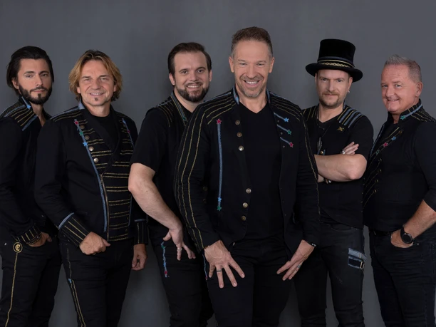 Höhner Six smiling men in black clothing and festive designs pose in front of a gray background.</p> <p
