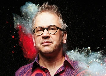 Robert Griess Porträt eines Mannes mit Brille vor schwarzem Hintergrund, umgeben von buntem Farbpulver.