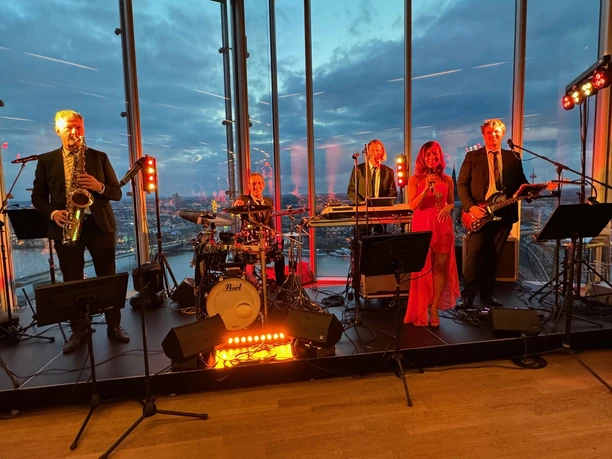 Confetti & Friends Eine sechsköpfige Band spielt vor einer gläsernen Skyline bei Sonnenuntergang.