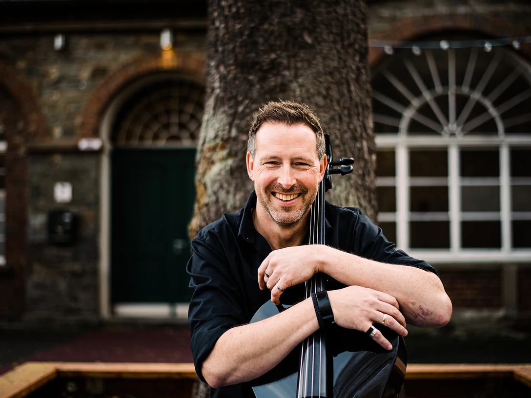 Simply Cello Mann lächelt, das Cello entspannend halten vor historischer Backsteingebäude-Fassade.Man smiling, holding the cello relaxing in front of a historic brick building façade.