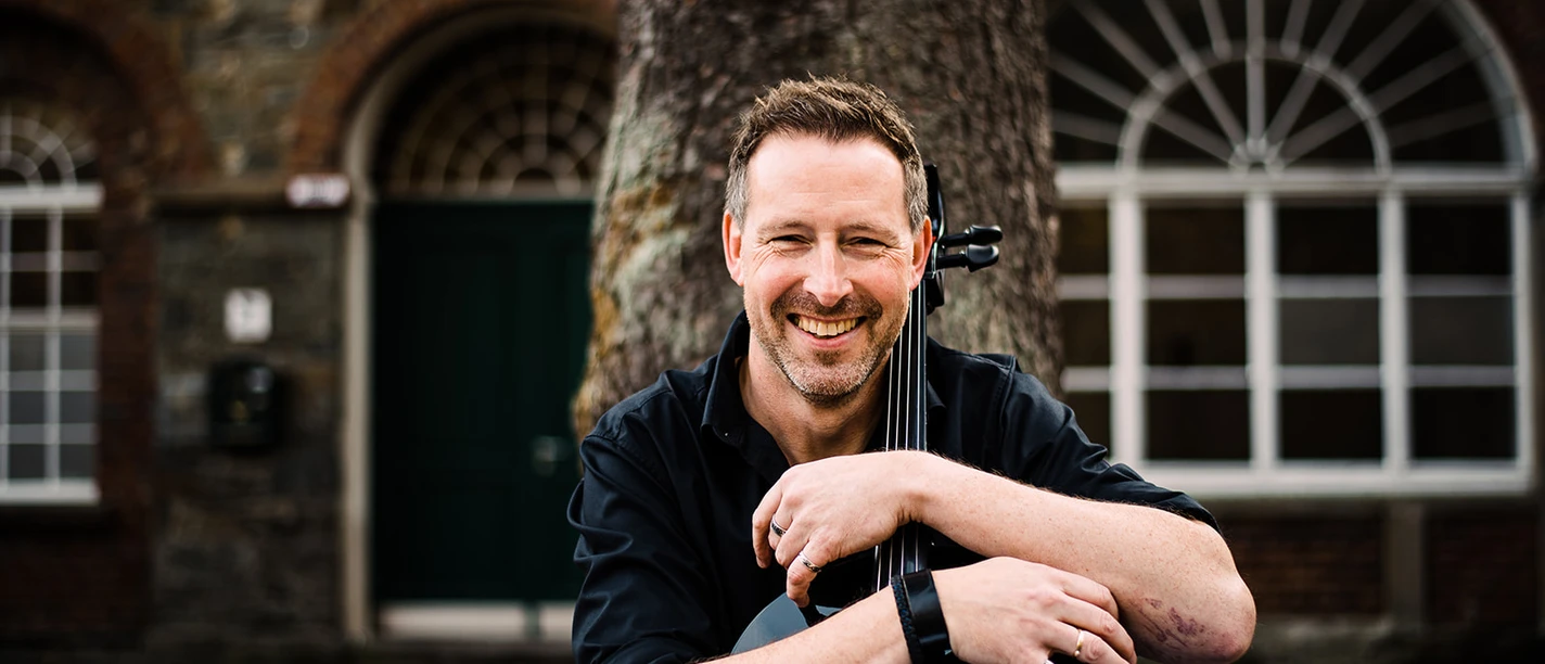 Simply Cello Mann lächelt, das Cello entspannend halten vor historischer Backsteingebäude-Fassade.