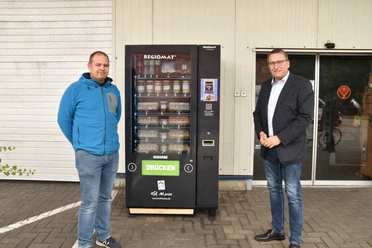 © Gemeinde Salzbergen Zwei Männer stehen vor einem Verkaufsautomaten mit regionalen Lebensmitteln auf einem Hof.