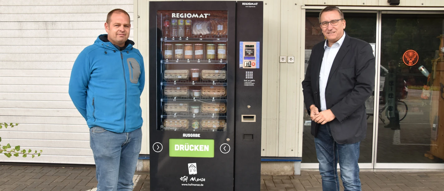 © Gemeinde Salzbergen Zwei Männer stehen vor einem Verkaufsautomaten mit regionalen Lebensmitteln auf einem Hof.