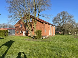 © Gemeinde Salzbergen Backsteingebäude mit rotem Dach auf grüner Wiese, umgeben von Bäumen unter blauem Himmel.