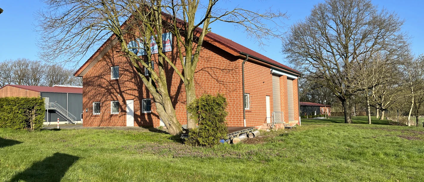 © Gemeinde Salzbergen Backsteingebäude mit rotem Dach auf grüner Wiese, umgeben von Bäumen unter blauem Himmel.