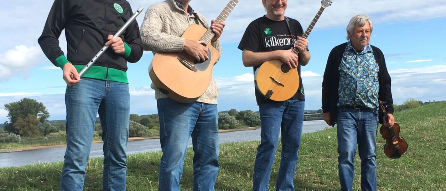 Kilkenny Msik.jpg Vier Musiker mit Instrumenten stehen im Freien vor einer Flusslandschaft und lachen in die Kamera.