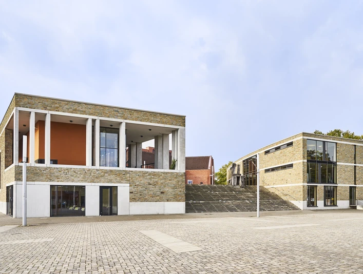 Kulturzentrum_Aussen_047.jpg Modernes Gebäude mit Glasfronten und Backsteinmauern auf einem gepflasterten Platz bei klarem Himmel.</p>Modern building with glass fronts and brick walls on a paved square under a clear sky.</p> <p