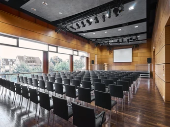 Kulturzentrum_kleiner_Saal_068.jpg Moderner Konferenzraum mit Holzwänden, leeren schwarzen Stühlen und einer großen Leinwand.</p>Modern conference room with wooden walls, empty black chairs and a large screen.</p> <p
