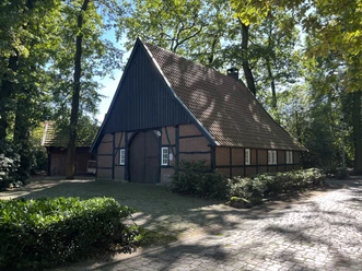 © Gemeinde Salzbergen Fachwerkhaus mit rotem Ziegeldach, umgeben von Bäumen und Kopfsteinpflaster im Sonnenschein