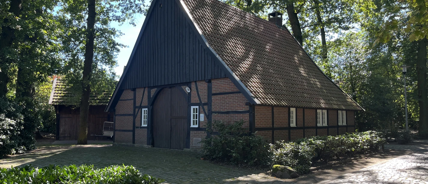 © Gemeinde Salzbergen Fachwerkhaus mit rotem Ziegeldach, umgeben von Bäumen und Kopfsteinpflaster im Sonnenschein