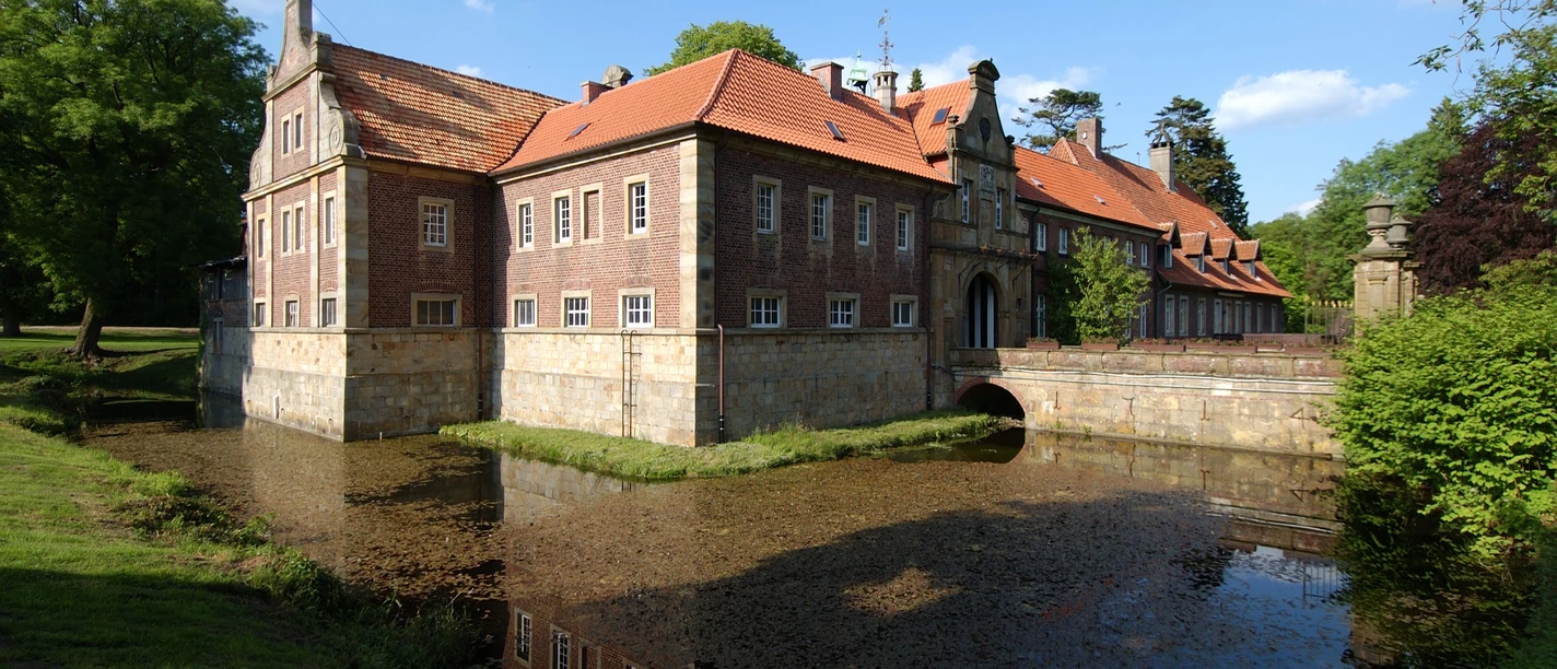 © Gemeinde Salzbergen Backsteinherrenhaus mit rotem Ziegeldach, Wassergraben und Brücke, umgeben von Bäumen und Wiese.