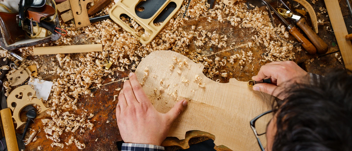 CC0_Erlebniswelt Musikinstrumentenbau_1_(c)_Danny-Otto (20).jpg Überblick auf Tisch in Werkstatt, Person schnitzt Geigenkörper aus hellem Holz.