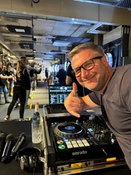 DJ Mr. Easy Ein DJ mit Brille vor einem Mischpult, das eine Veranstaltung mit Leuten im Hintergrund zeigt.</p>