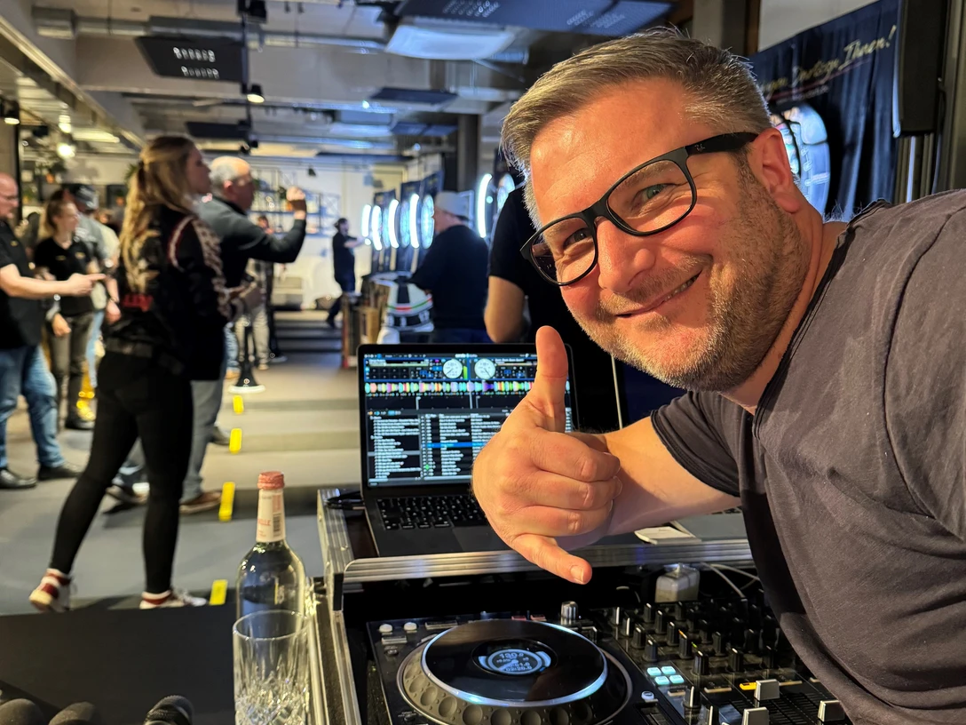 DJ Mr. Easy Ein DJ mit Brille vor einem Mischpult, das eine Veranstaltung mit Leuten im Hintergrund zeigt.</p>A DJ with glasses in front of a mixing desk showing an event with people in the background.</p> <p