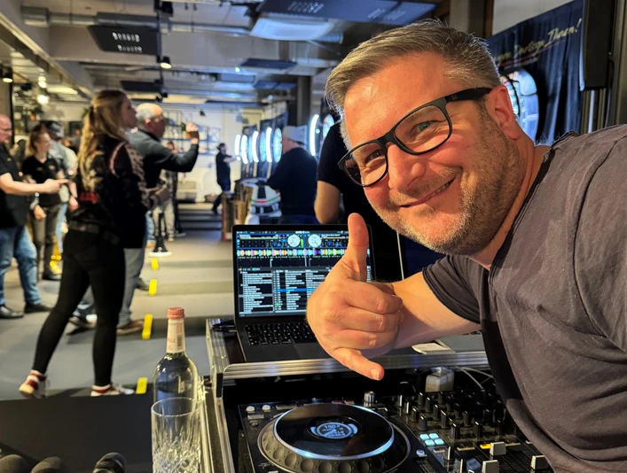 DJ Mr. Easy Ein DJ mit Brille vor einem Mischpult, das eine Veranstaltung mit Leuten im Hintergrund zeigt.</p>A DJ with glasses in front of a mixing desk showing an event with people in the background.</p> <p