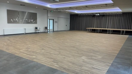 © Gemeinde Salzbergen Moderner Veranstaltungssaal mit Holzfußboden, grauen Vorhängen und heller LED-Deckenbeleuchtung.