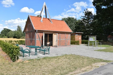 © Gemeinde Salzbergen Backsteinbau mit rotem Satteldach und Holzfachwerk, davor Sitzbänke und Infotafel im Grünen.