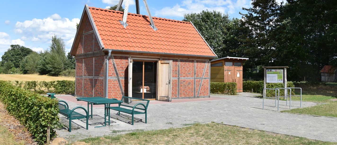 © Gemeinde Salzbergen Backsteinbau mit rotem Satteldach und Holzfachwerk, davor Sitzbänke und Infotafel im Grünen.