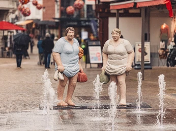 Zwei Statuen in bunten Badeanzügen stehen auf einem gepflasterten Platz neben Wasserfontänen.