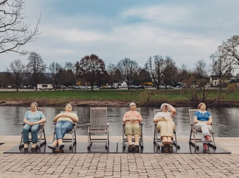 Fünf lebensgroße Figuren in bunten Alltagskleidern sitzen auf Liegestühlen an einem Flussufer.