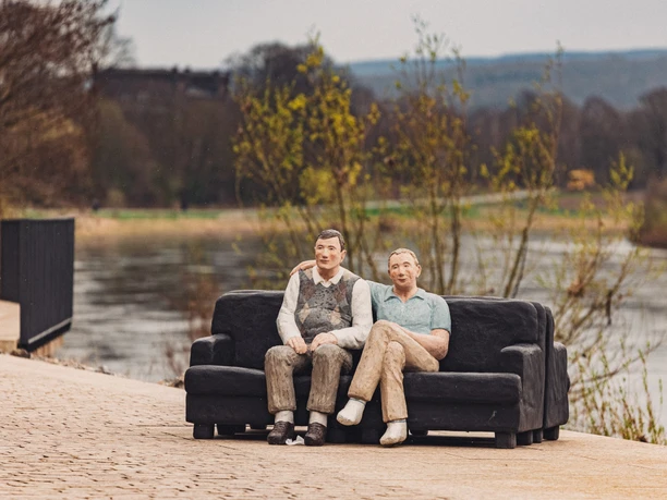 Zwei lebensgroße Figuren aus Holz sitzen auf einem Sofa an einem Flussufer in ländlicher Umgebung.
