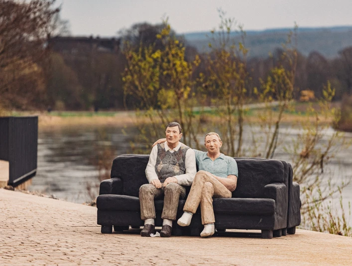 Zwei lebensgroße Figuren aus Holz sitzen auf einem Sofa an einem Flussufer in ländlicher Umgebung.