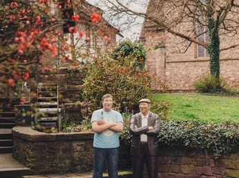 Zwei lebensnahe Skulpturen stehen in einem grünen Garten mit blühenden roten Blumen und Backsteingebäude.
