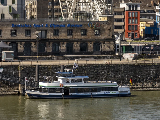 Kölntourist passenger boat service The picture shows a large white Ferris wheel, prominently enthroned in the skyline of Cologne on the banks of the Rhine in the background. An excursion boat on the Rhine can be seen in the foreground, while the cityscape is dominated by historic brick buildings.