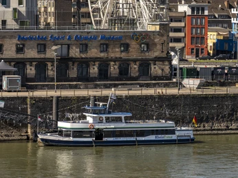 Kölntourist Personenschiffahrt Das Bild zeigt ein großes weißes Riesenrad, das prominent in der Skyline von Köln am Rhend im Hintergrund histinufer thront. Im Vordergrund ist ein Ausflugsschiff auf dem Rhein zu sehen, währeorische Backsteingebäude das Stadtbild prägen.The picture shows a large white Ferris wheel, prominently enthroned in the skyline of Cologne on the banks of the Rhine in the background. An excursion boat on the Rhine can be seen in the foreground, while the cityscape is dominated by historic brick buildings.
