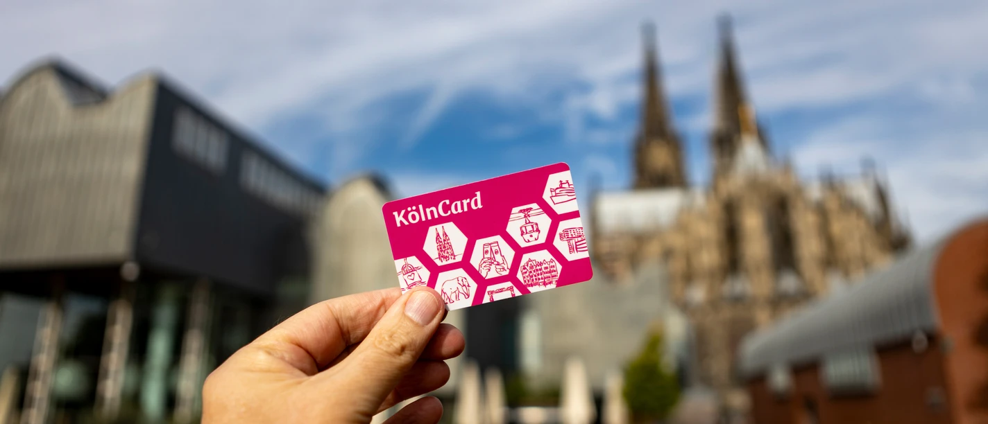 KoelnCard-bearb-KoelnTourismus-Seelbach-4142.jpg KölnCard in Hand vor dem unscharfen Kölner Dom und modernem Gebäude im Hintergrund.