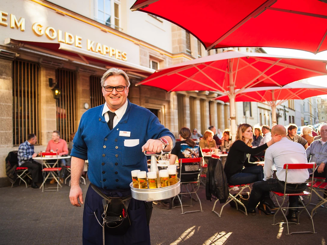Traditioneller Köbes <p>Ein lächelnder Kellner serviert Bier unter roten Sonnenschirmen in einem belebten Außenbereich.</p><p>A smiling waiter serves beer under red parasols in a lively outdoor area.</p>