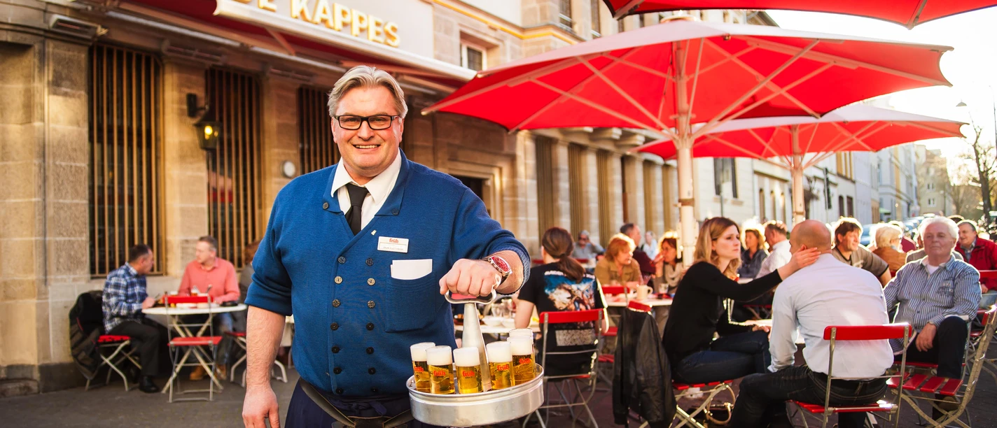 Traditioneller Köbes <p>Ein lächelnder Kellner serviert Bier unter roten Sonnenschirmen in einem belebten Außenbereich.</p>