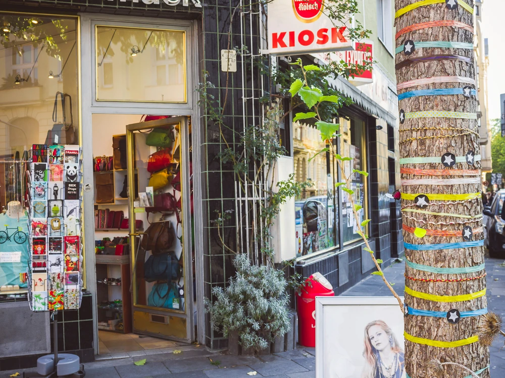 Belgisches Viertel Köln <p>Ein Kiosk in einer städtischen Straße, dekoriert mit bunten Taschen und einem kunstvoll verzierten Baum.</p><p>A kiosk in an urban street, decorated with colorful bags and an artistically decorated tree.</p>
