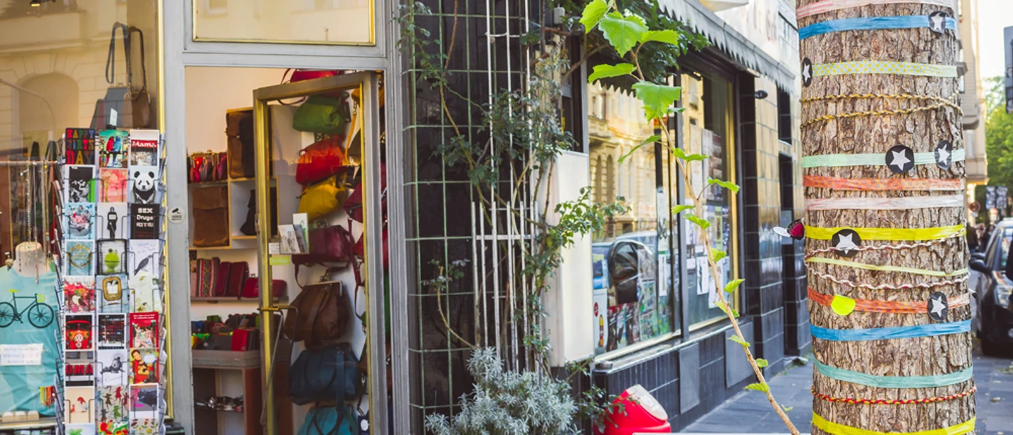 Belgisches Viertel Köln <p>Ein Kiosk in einer städtischen Straße, dekoriert mit bunten Taschen und einem kunstvoll verzierten Baum.</p>