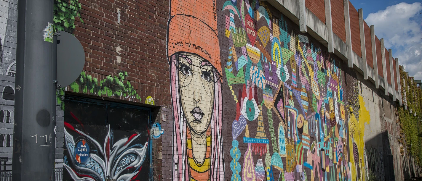 Street Art in Köln Ehrenfeld <p>Buntes Graffiti-Kunstwerk auf roter Backsteinwand zeigt Porträt und abstrakte Formen in leuchtenden Farben.</p>