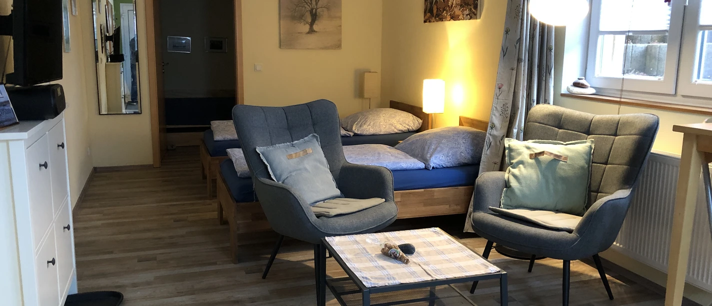 Apartments Am Kaldenberg_Claudia Apsel-Nentwig.JPG Ein modernes Apartment mit zwei blauen Betten, grauen Sesseln und einem Tisch in einem hellen Raum.