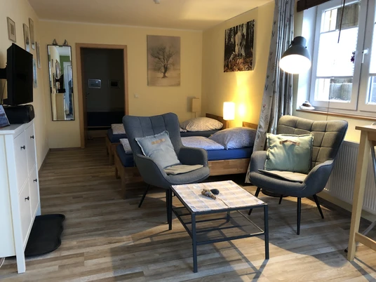 Apartments Am Kaldenberg_Claudia Apsel-Nentwig.JPG Ein modernes Apartment mit zwei blauen Betten, grauen Sesseln und einem Tisch in einem hellen Raum.