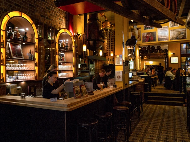 Hellers_Brauhaus_Titel Die Bar im warmen Licht, mit Holzmöbeln und Gästen, strahlt gemütliche Atmosphäre aus.