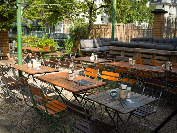 brauwelt-biergarten-22.jpg An idyllic beer garden with wooden tables and chairs in the open air, surrounded by green vegetation.