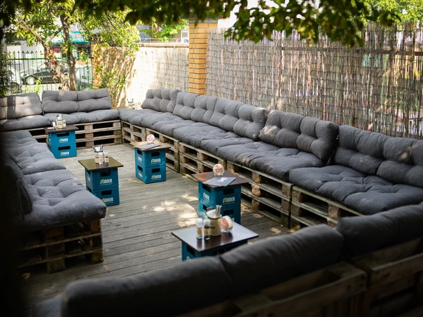 brauwelt-biergarten-24.jpg <p>Gemütliche Lounge mit grauen Polstern auf Holzpaletten in einem sonnigen Hinterhof.</p>