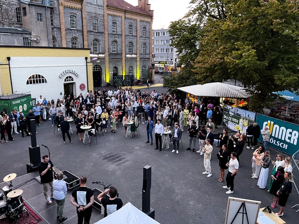 IMG_2236.jpg <p>Menschenmenge auf einem belebten Freiluft-Event mit Bühne, Getränkeständen und historischen Gebäuden.</p>