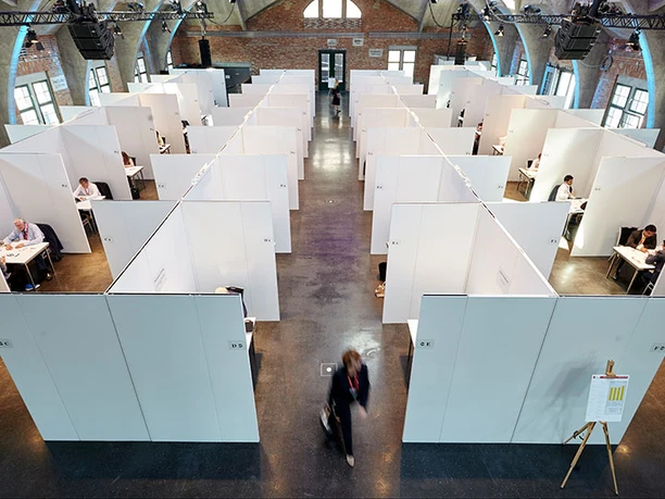 Innenansicht einer großen Messehalle mit weiß geteilten Kabinen und Menschen bei der Arbeit.