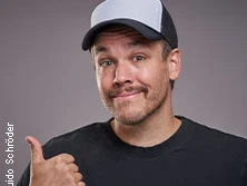 Ein Mann mit Baseballkappe und schwarzem T-Shirt lächelt, hebt den Daumen und blickt zur Kamera.