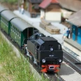 Eisenbahnwelten Rathen Modelleisenbahn Modelleisenbahn mit schwarzer Dampflok und grünen Waggons auf Gleisen, umgeben von Miniaturgebäuden und Figuren, bei sonnigem Wetter.