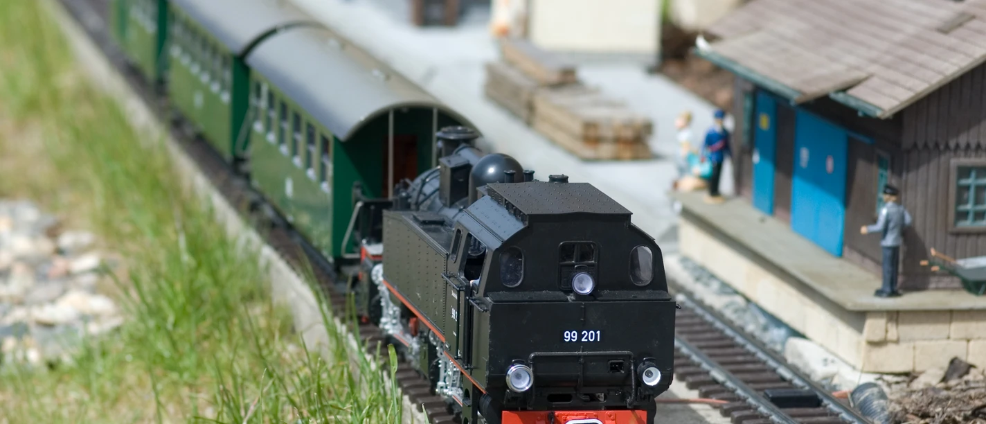 Eisenbahnwelten Rathen Modelleisenbahn Modelleisenbahn mit schwarzer Dampflok und grünen Waggons auf Gleisen, umgeben von Miniaturgebäuden und Figuren, bei sonnigem Wetter.