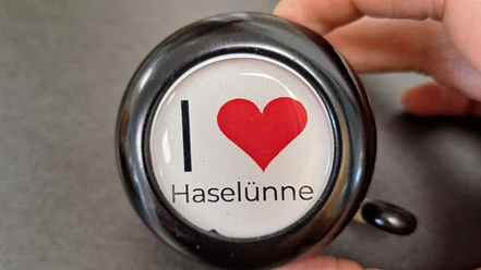 Fahrradklingel Haselünne Schwarze Fahrradklingel mit Aufdruck I Love Haselünne und rotem Herzsymbol auf grauem Hintergrund.