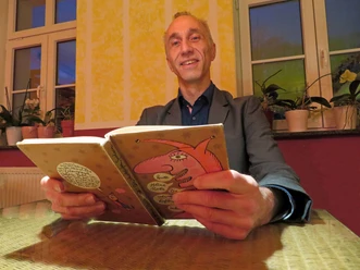 Uwe Becker Uwe Becker liest lächelnd in einem gemütlichen Raum vor einem Hintergrund aus gelben Wänden.