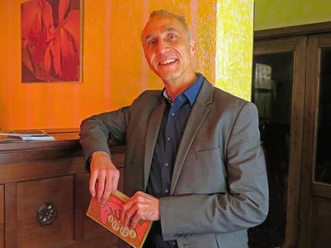 Een man van middelbare leeftijd in een grijs pak houdt een boek vast en lacht vriendelijk in de camera.
