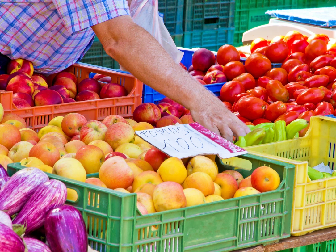 Wochenmarkt (1).png Frischwarenstand auf einem Markt zeigt bunte Früchte wie Äpfel, Tomaten und Paprika in Kisten.