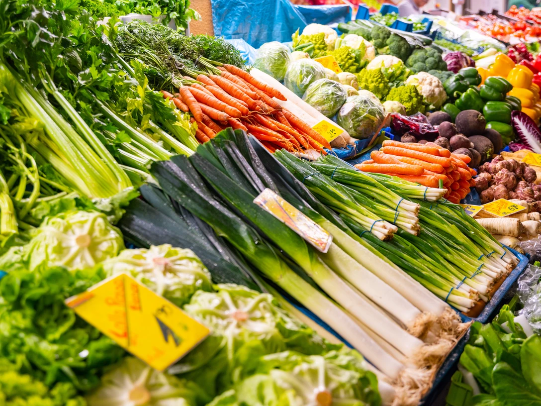 Wochenmarkt (2).png Frisches Gemüse in leuchtenden Farben, darunter Karotten, Lauch und Kohl, auf einem Wochenmarktstand.